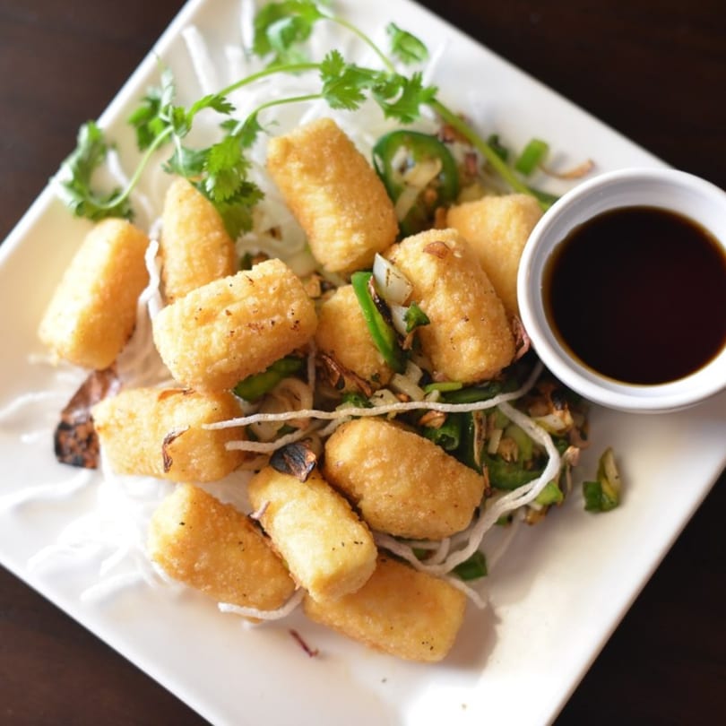 Crispy fried tofu pieces with cilantro, jalapeños, and a side of soy sauce on a white plate.