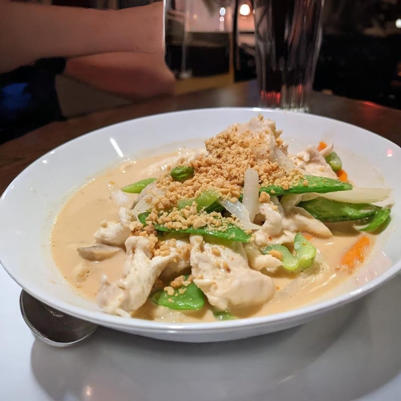 Asian chicken curry with snow peas, carrots, and crushed peanuts in a white bowl.