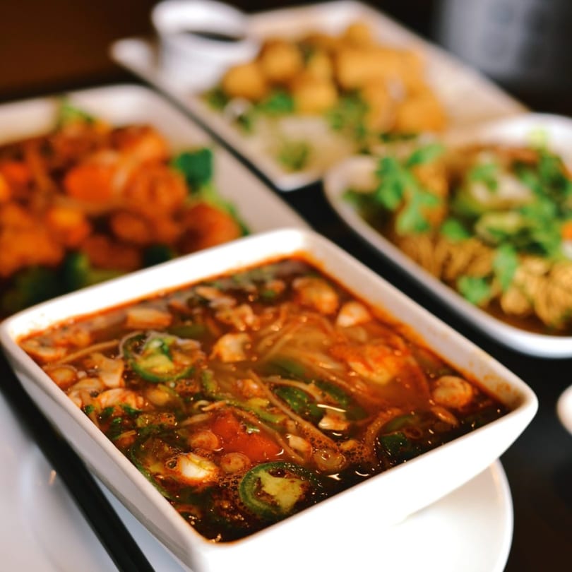 Spicy Asian soup with jalapenos and mushrooms, with other Asian dishes in the background.